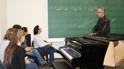 Eine Gruppe Menschen schaut auf eine Tafel, auf der musikalsiche Noten aufgeschrieben sind. Ein Mann erklärt.